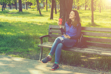 Beautiful Caucasian woman with long brown hair sitting on a yoga mat in the park drinking a cup of tea or coffee and smiling. Middle-aged athletic woman with long hair drinks coffee in a park. tonedの写真素材