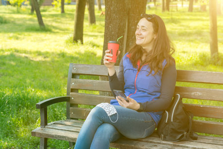 Beautiful Caucasian woman with long brown hair sitting on a yoga mat in the park drinking a cup of tea or coffee and smiling. Middle-aged athletic woman with long hair drinks coffee in a parkの写真素材