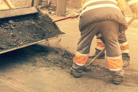 Road workers in bright orange reflective uniforms use shovels to scrape accumulated sand. maintenance of road and highway pavements. pothole repairing works. tonedの写真素材