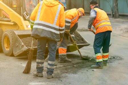 Road workers in bright orange reflective uniforms use shovels to scrape accumulated sand. maintenance of road and highway pavements. pothole repairing works. tonedの写真素材