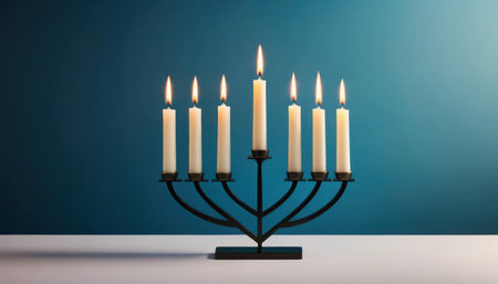 A Hanukkiah holds nine candles, all lit, set on a flat surface with a blue wall behind it.の素材