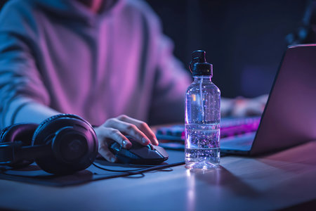 A gamer is hydrating during a session while using a mouse and headphones set in neon lighting.の写真素材