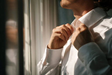 A man in a white dress shirt is buttoning his collar in front of a window with sunlight streaming...