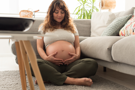 Happy pregnant woman sitting and touching her belly at homeの写真素材