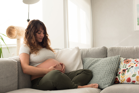 Happy pregnant woman sitting and touching her belly at homeの写真素材