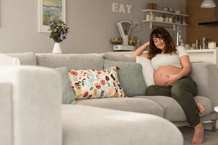 Happy pregnant woman sitting and touching her belly at homeの写真素材
