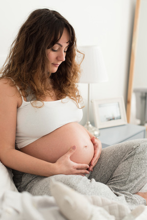 Happy pregnant woman sitting and touching her belly at homeの写真素材