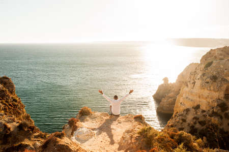 Man sitting on the rocks with open arms to the ocean view - Sunset lightの写真素材