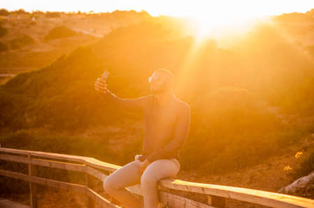 Latin man taking selfies with a smartphone over an amazing sunset landscape view. Natural  outdoor lightの写真素材