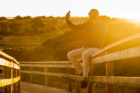 Latin man taking selfies with a smartphone over an amazing sunset landscape view. Natural  outdoor lightの写真素材