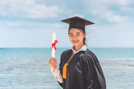 Graduated woman in cap and gown holding certificated celebrating in Commencement day with blue sky on the beach with beautiful sea in summer so proud and happiness,Education Success in summer Conceptの写真素材