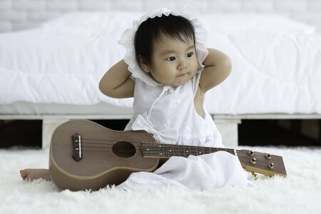 Cute Sweet Adorable Asian Baby wearing white dress sitting with guitar on Carpet smiling and playing with happiness emotional in cozy bedroom,Healthy Baby Conceptの写真素材