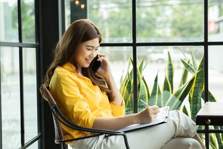 Beautiful Attractive Asian Business woman writing down requirement to book and talking with customer in smartphone,Feeling so happiness and working with service mind,Business and Finance Conceptの写真素材