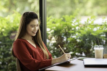 Beautiful Attractive Asian woman wearing red dress working with laptop smile and writing idea and...