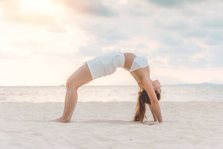 Asian young woman practice Yoga bridge Pose on the sand and beach with sunset beautiful sea in...