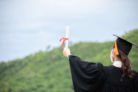 back of view Graduate put hands up  celebrating wear Medical mask for protection virus holding diploma or certificated due Virus outbrake and COVID-19 virus flu pandemic,New normal of graduation dayの写真素材