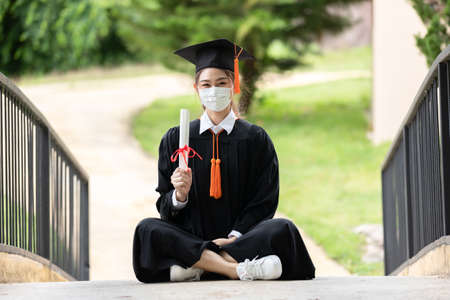 Graduated woman wear cap gown and  Medical mask for protection virus holding diploma or certificated due Virus outbrake and COVID-19 virus flu pandemic,New normal of graduation dayの写真素材