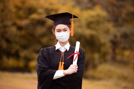 Graduated woman wear cap gown and  Medical mask for protection virus holding diploma or certificated due Virus outbrake and COVID-19 virus flu pandemic,New normal of graduation dayの写真素材