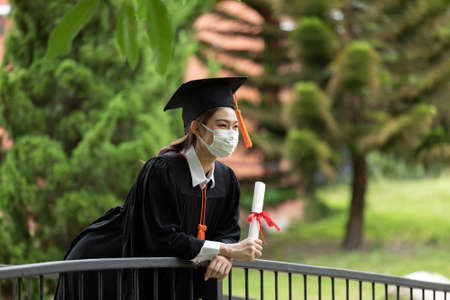 Graduated woman wear cap gown and  Medical mask for protection virus holding diploma or certificated due Virus outbrake and COVID-19 virus flu pandemic,New normal of graduation dayの写真素材