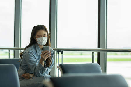 Asian young woman traveller wearing face maks using mobile phone at airport Due Covid-19 flu virus pandemic and protectionの写真素材