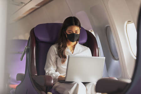 Asian young woman wearing protective mask using laptop sitting near windows at first class on airplane during flight,Traveling and Business during COVID-19 virus pandemic conceptの写真素材