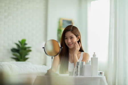 Happy Asian young woman smile and looking in mirror doing skin care touch cheek and beauty treatment at home,Wellness woman self care with beauty product at homeの写真素材