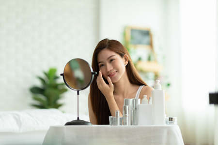 Happy Asian young woman smile and looking in mirror doing skin care touch cheek and beauty treatment at home,Wellness woman self care with beauty product at homeの写真素材