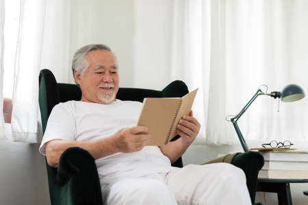 Asian Elderly man with white hairs sitting on sofa reading book ralax and cozy at homeの写真素材