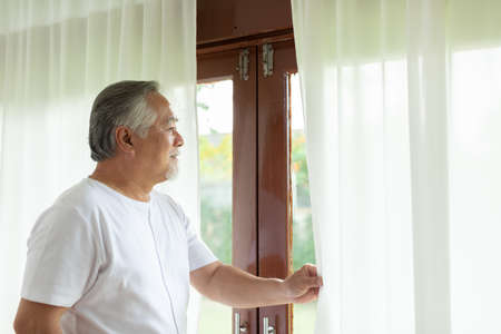 Happy Elder Asian man looking outside window in the morning smile and relax at homeの写真素材