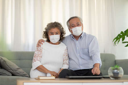 Happy Asian Elderly or Seniors couple wearing medical mask to prevent  sitting on sofa in living room at home,New normal lifestyle of older stay at homeの写真素材