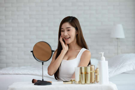 Happy Wellness Asian young woman smile and looking in mirror doing skin care touch cheek massage and beauty treatment in cozy bedroom,Wellness preety girl self care with beauty product at homeの写真素材