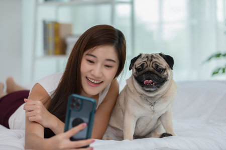 Beautiful Asian young woman take a photo selfie bu mobile phone with her cute dog pug breed in cozy bedroom,Happiness girl playing with dog to take a rest at home in holidayの写真素材