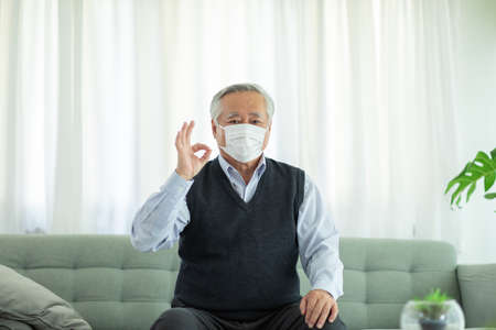 Happy Asian Elderly or Seniors man wearing medical face mask to prevent coronavirus making OK after received a coronavirus vaccine sit on sofa in cozy living room at homeの写真素材