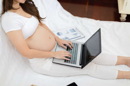 Pregnant business woman sitting and working on the laptop with sale report,tablet,books and sticky note at home,Pregnant of businesswoman typing on laptop,Business and Pregnancy Conceptの写真素材