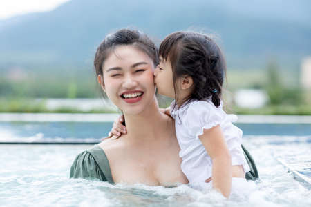 Adorable kid girl kissing her mother  with love and happiness feeling in swimming pool.Asian young mother smile with her sweet little girl kiss on her cheek.Love and family conceptの写真素材