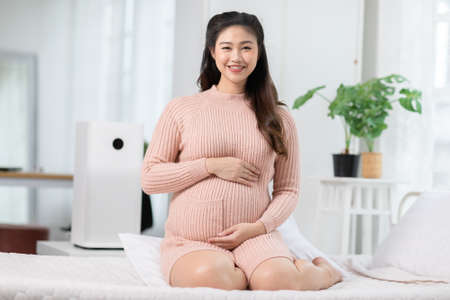 Happy Pregnant Asian Woman sitting on bed holding and stroking her big belly at home.Pregnancy of...