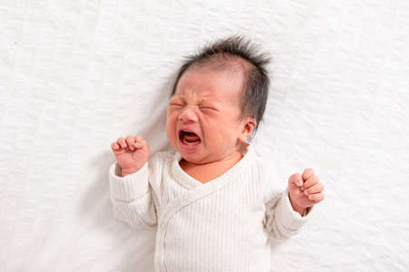 Crying Newborn baby boy lying on white bed at home.Infant baby screaming very hungry or stomach pain.Tired baby crying conceptの写真素材