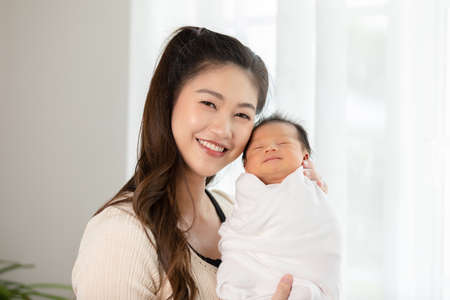 Beautiful Asian woman holding newborn baby in her arms standing in front of windows at cozy home.Happy infant baby sleep in mother arms safety and comfortable.Mom and Baby Conceptの写真素材