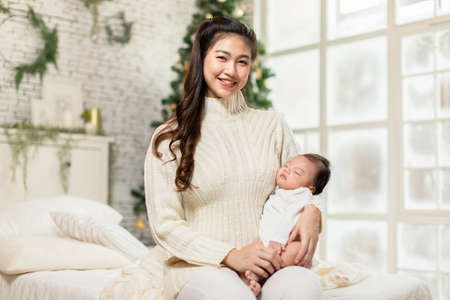 Beautiful Asian woman holding newborn baby in arms sit on bed at white cozy home with christmas tree...