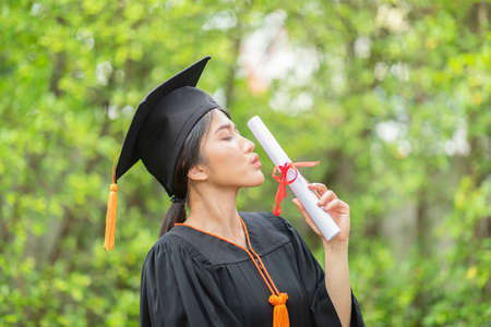 Attractive Asian Women Student Graduate in cap and gown celebrating kissing certificate in hand and...