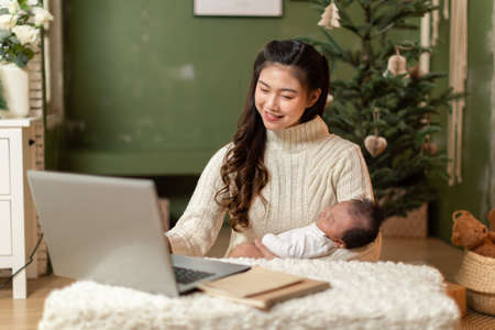 Happy Asian mother woman holding her baby newborn and working staying at home.Freelance mom working...