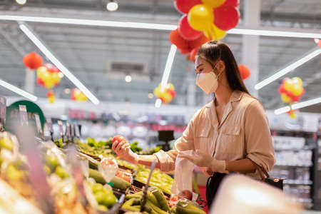 Beautiful asian woman wearing medical face mask and rubber gloves choosing tomato on vegetable and fruits shelf in supermarket.Female looking and hold tomato shopping in grocery department storeの写真素材