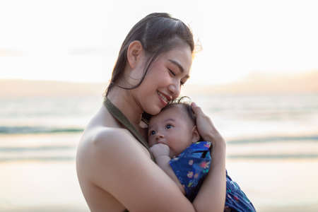 Beautiful Attractive Asian mom holding her baby newborn in hand and hugging baby sweet and lovely outdoor on the beach.Happy mother and infant baby looking together smile with natural sunsetの写真素材