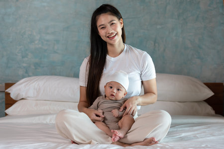 Beautiful Asian mom woman holding newborn baby in her arms sit on bed at white cozy home.Happy...