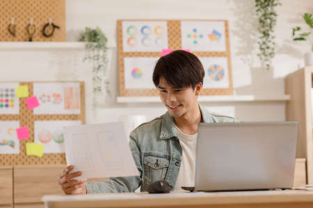 Entrepreneur handsome business asian man wear casual shirt working with laptop and look at document to compare something at home.Happy Attractive Freelance smart working online enjoy with own businessの写真素材