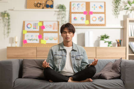 Clam of Asian young man doing yoga lotus pose to meditation and relax on couch during work online at home.Happiness male break after worked close him eyes and deep breath with yoga so peaceful.の写真素材
