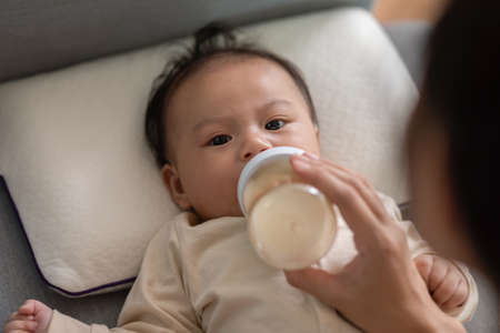 Close up hand of mother holding milk bottle Newborn baby lying on bed drinking milk.Cute infant baby feeding milk with love at home.Newborn Baby Conceptの写真素材