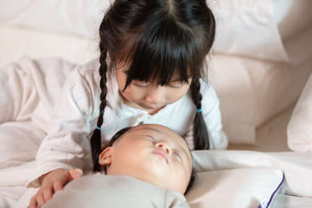 Cute Sister looking at brother sleep on bed with love.Infant newborn baby sleep on bed and sister stay beside to take care of young brother siblings happiness and comfortable at cozy home.Good momentの写真素材