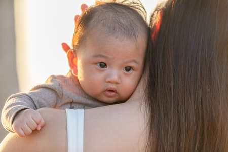 Beautiful Attractive Asian mom holding her baby newborn in hand and kissing on baby head sweet and lovely outdoor on the beach.Happy mother and infant baby looking together smile with natural sunsetの写真素材