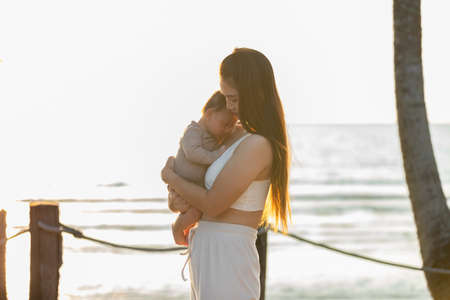 Sleeping baby boy is snuggling with mom is the most warmest moment. An innocent Asian kid is chubby, cute in grey pajamas. Asian mom holding his son at beach front while the sunset.の写真素材